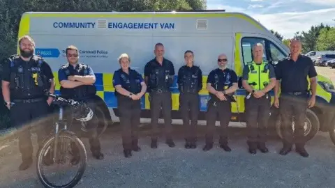 Eight police officers stand in front of a police van branded with Community Engagement Van. There is also a bicycle in the shot in front of a male officer. 