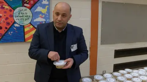 A man in a dark suit is in the image.  He is standing in a school hall, there is a colourful piece of childrens' artwork behind him.  The man is holding a small tub, which contains a pudding called Noah's pudding.  To the right of him is a table set up with many more tubs of the pudding.