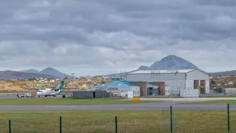Getty Images Carrickfinn Airport in County Donegal. An Aer Lingus plane sits on the runway near the airport, a small terminal building with a blue roof is to the right and beside that is a large hangar. Mountains can be seen in the background.