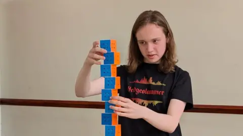Louise Allinson Thomas, who is has shoulder-length hair and a black t-shirt is standing in front of a pile of cubes.