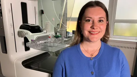 Rebecca Corbett, Quality and Improvement Manager for West Sussex Brighton and Hove Breast screening Service. Rebecca, who is sat in front of the screening machine, is wearing a blue t-shirt and has her hair down. 