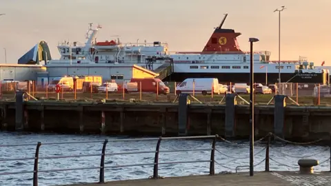 A large passenger ferry docked in a harbour. Parked vehicles can be seen in the foreground