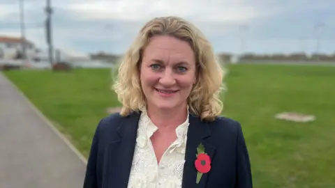 Louise McKinlay stands on a pathway in front of a section of green grass. She has shoulder length blonde hair that is slightly curled. She wears a navy suit jacket with a poppy on one side of her chest and a cream blouse underneath.