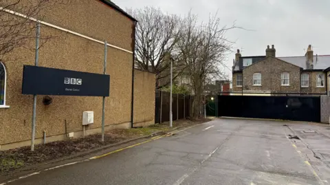 Danny Fullbrook/BBC The access road that runs towards the EastEnders set. A large black sign reads "BBC Elstree Centre". Houses can be seen on the other side of a black security gate