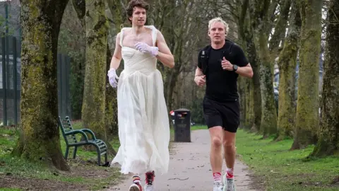 Getty Images BBC Radio 1 presenter Jamie Laing is wearing a black t-shirt and shorts and is running alongside Greg James in a white wedding dress, through a park