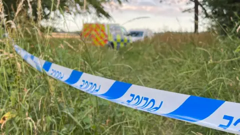 Stuart Woodward/BBC Two police vans parked on a field, beyond blue and white police tape.