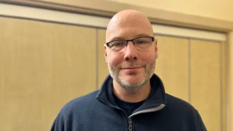 A bald man with a grey beard wearing a navy zip up top smiling at the camera
