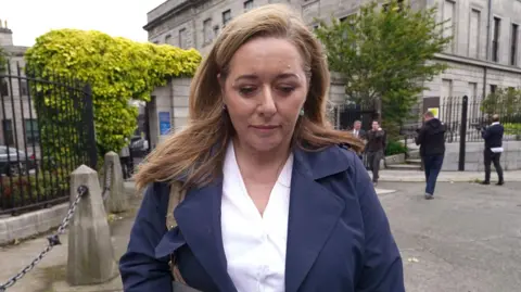 Brian Lawless/PA Wire BBC Spotlight NI reporter Jennifer O'Leary outside the High Court in Dublin.  She has long, straight auburn hair and is wearing a navy trench coat over a white blouse. 