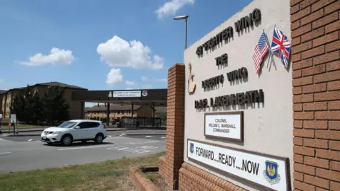 Getty Images One of the entrance gates into RAF Lakenheath. A large sign detailing the name of the base with flags on it rests to the side of the entrance. Ahead a security gate can be seen with a white car travelling out. 