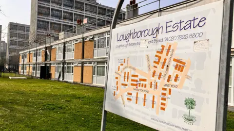 A general view of the outside of an estate. There is a two-storey section with grass in front of the building at the forefront of the image. Behind are two higher blocks. There is a map of the site with the words Loughborough Estate written across it. 