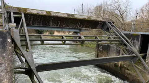 An iron brown gate with water flowing beneath and trees in the background.