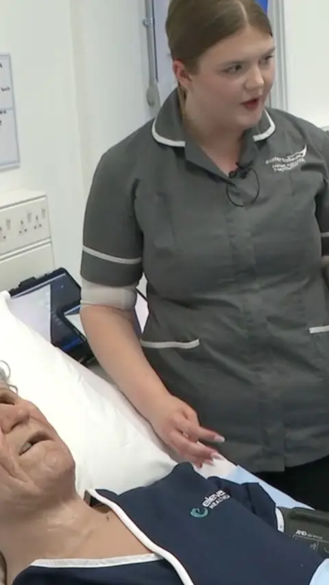 A lifelike patient mannequin on a bed. There is a woman in grey nursing uniform next to the bed.