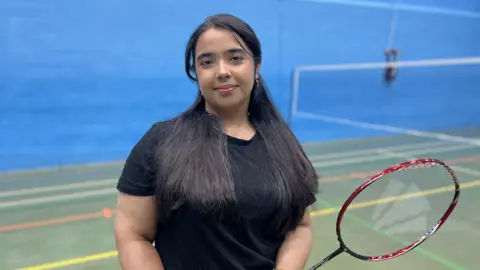 A young girl with long black hair smiles into the camera. She is wearing a black short sleeved top and holding a red badminton racket. Behind her is a sports hall with a badminton net, a green floor and blue walls. 