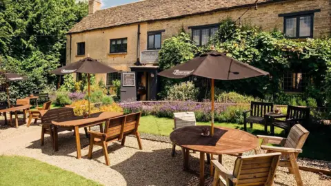 The Bell at Sapperton A photo showing a pub garden with a Cotswold stone building in the background. It shows a number of tables with umbrellas and chair around them