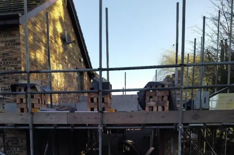 David Eddon An image of a house with a half built extension on its right hand side. The extension is one storey tall with scaffolding all over and piles of bricks on top ready for the next level. 