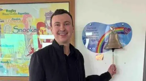 Man with dark brown short hair wears an all black outfit and is smiling - Sam Birchall. The 24-year-old stands in a hospital room in front of a bronze bell on a rainbow mount. He holds the string of the bell as if to begin ringing it.