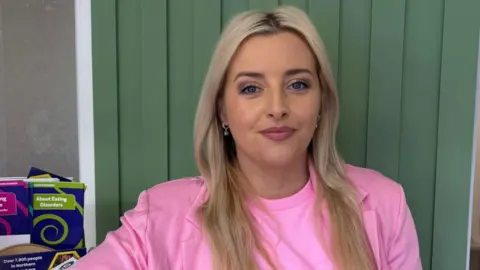 Órlaithí Flynn has blonde hair, a side parting and gold earrings. She is wearing a 
bright pink t shirt and a pink jacket and is standing in front of a wood-panelled wall which is painted in green