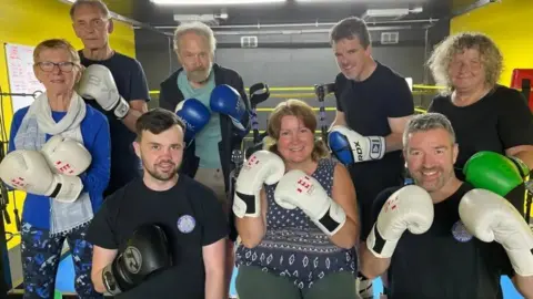 Members of the Parky Blinders project in a boxing gym. They are all wearing boxing gloves and looking at the camera. A boxing ring is visible in the background, surrounded by bright yellow walls.