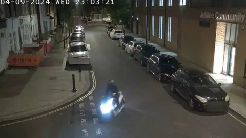 Metropolitan Police handout A screen shot from CCTV of a motorcyclist turning left at a side street junction, opposite a building with the words Jubilee Community just seen above a doorway.
There are parked cars on either side of the road but no pedestrians are visible.