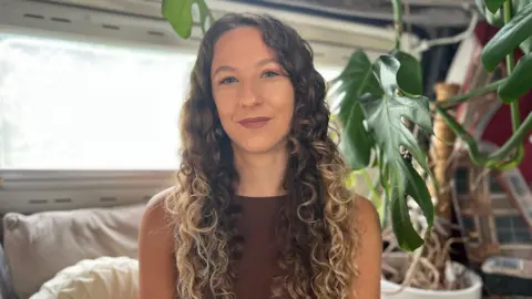 Sophie Robert pictured with long, curly brunette hair that turns blonde towards the end sitting indoors beside a large leafy houseplant, wearing a sleeveless brown top.