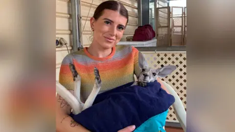 Rosie Scott Rosie Scott, fotografada segurando um bebê canguru. O canguru está enrolado em uma toalha azul marinho e segura uma pata na ponta da toalha. Rosie está vestindo uma camiseta gradiente laranja e verde de manga curta e sorri para a câmera. Ela tem cabelos castanhos presos para trás e olhos castanhos. 