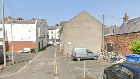 A narrow through road flanked by terraced properties. Small car parks on either side of the road are in the foreground and a load of beer barrels and what appears to be fly-tipped waste are in one of the car parks.