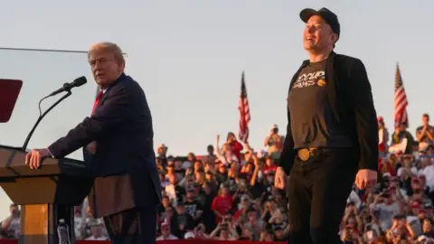 Getty Images Donald Trump, in a navy suit and red tie, stands at a podium, looking sideways at the camera while Elon Musk, dressed in black, beams before a crowd a with American flags in the background