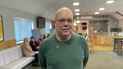 Mark Ansell / BBC A middle-aged man with bald head and wearing metal-rimmed glasses and green crew-necked top stands in a room with several people seated in the background.