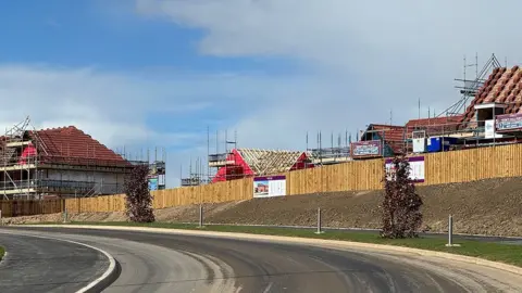 Four houses are being constructed on a building site and are all still covered by scaffording behind a wooden fence. In front of the buildings is a newly laid road with a wide pavement 