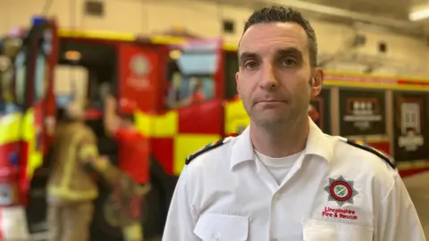A man with short brown hair that is spiked up pulling a serious expression and looking into the camera. He is wearing a white shirt that says 'Lincolnshire Fire & Rescue' on it with a star logo. The background is blurred but there is a red and yellow fire truck with the doors open that you can see a couple of people standing next to.