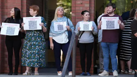 PA Four women and a man are standing in front of a brick building holding small white posters that say 'regulate our funeral industry'