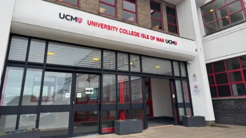 The entrance to the UCM building, which has brown bricks and features white concrete cladding and pillars. The entrance is glass-fronted and has UCM University College Isle of Man written in black and red above it.