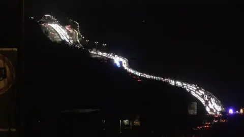 A night time scene showing a long bendy road with a queue of traffic, with white car headlights lined up. On the right edge of the picture blue flashing lights can just be seen.