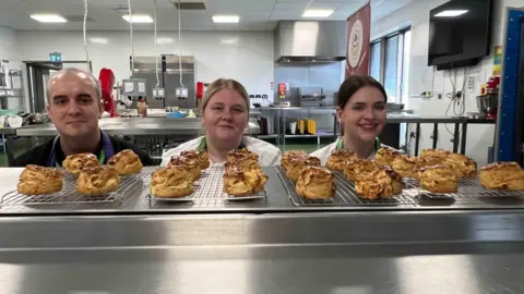 Kate Bradbrook/BBC A man and two women are in a kitchen standing in front of trays of baked scones, which sit in the foreground.