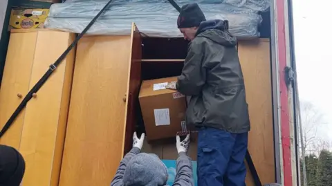 Help Ukraine BAMK UK Community Interest Company (CIC) Two people loading up a lorry that is full of wardrobes. They are putting a box into the wardrobe. One man is wearing jeans, a dark hat, dark coat and is side on. The other man has his arms out, wearing grey gloves, a grey hoodie, with the hood up. The doors are open and there are trees to the right. 