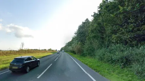 The A road is bordered by dense, green forest on one side and a green field and hedgerows on the other. There are a few cars, with their number plates blurred out, on the single carriageway road.