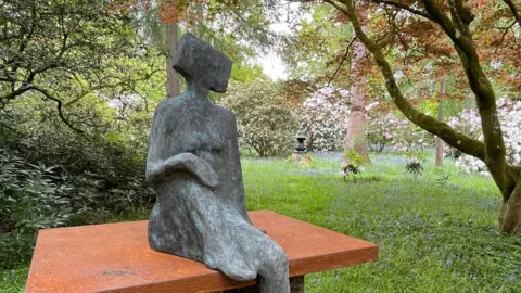 Ramster Garden A grey statue of a faceless person with a square head, sat on a terracotta ledge. The statue is situated in a green garden.
