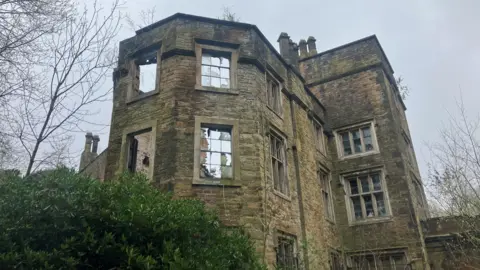 Photograph of the derelict Winstanley Hall in Wigan. The image shows smashed windows. The roof has partially collapsed.