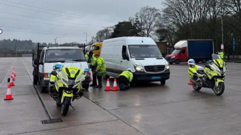 A van and flat-bed truck, both white, are pulled up between traffic cones on a stretch of concrete. Two police motorcycles are standing in front of them, while several people in hi-viz examine the vehicles.