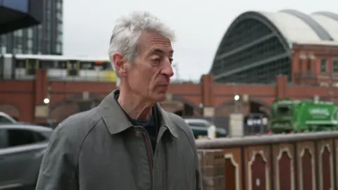 Andy Spinoza is standing in the centre of Manchester in front of a tram line.