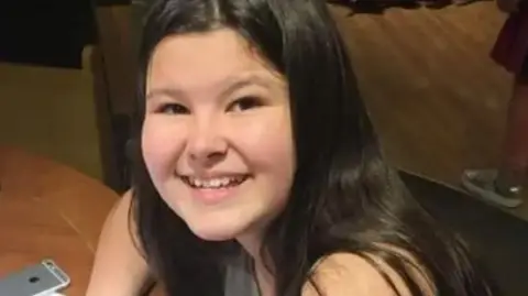 Jayne Renolds Leah Bedford sitting at a table. She is smiling and has long, black hair. A silver iPhone can be seen on the brown table.