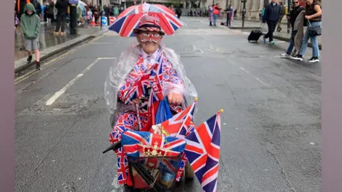 Tracey Joy A man riding a mobility scooter covered in Union flags. He has a small umbrella attached to his hat, which also has a Union flag design.