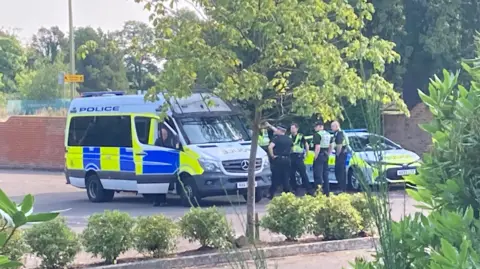 A police van and car in the hotel's car park with a group of four officers standing by the vehicles on 1 August 2024.