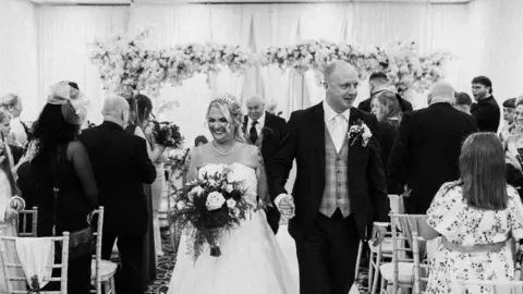 Lisa Marsay Lisa Marsay smiles in a wedding dress while holding her husband's hand and walking down the aisle at her wedding. 