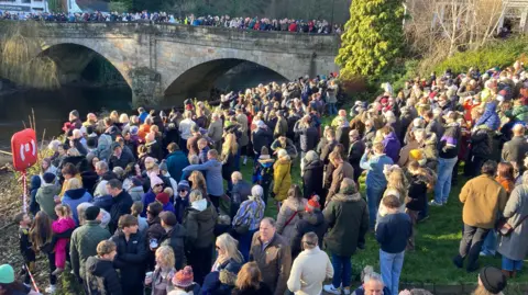 Cathy Killick/BBC A large outdoor gathering near a stone bridge with two arches spanning a river. The crowd is densely packed on the grassy riverbank in the foreground, with many people wearing winter coats, hats, and scarves, suggesting cold weather. Some individuals are holding drinks, and a few are looking at their phones. In the background, the bridge is lined with more people standing shoulder to shoulder, observing the scene below. Beyond the bridge, there are trees without leaves, indicating a winter season, and a white building with a sloped roof partially visible on the left side. A bright red life preserver is mounted near the riverbank.