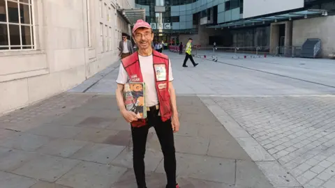 George Anderson stands in front of the BBC building holding a copy of Big Issue. He wears a red baseball cap and Big Issue gillet, and is smiling.