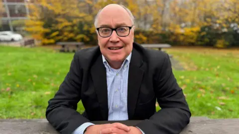 Prof Gary MacFarlane, a grey-haired man with glasses, wears a black suit and pale blue checked shirt, sitting at an outdoor table with grass and a yellow-leafed bush behind him.