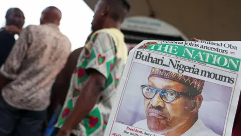 Reuters A person displays a newspaper carrying a headline reporting Muhammadu Buhari's death. Three men appear in the background of the image, out of focus. 