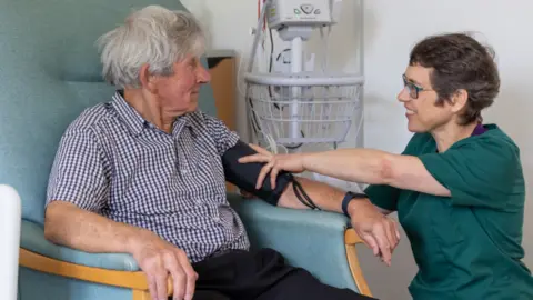 ReMind UK The picture shows a man and a nurse in a hospital setting.  The man is sitting in a chair having his blood pressure taken.