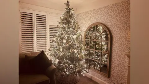 Joanne Kinsey A Christmas tree is decorated with silver baubles and white lights in the corner of a living room next to a sofa and mirror.